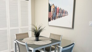 Un comedor con una mesa dispuesta para cuatro personas, rodeada de sillas blancas, una planta en maceta y un cuadro con chozas de playa en la pared.