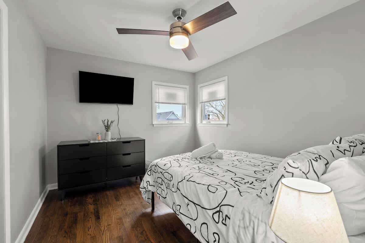 A bedroom with a bed, a ceiling fan, a TV mounted on the wall, and a dresser.