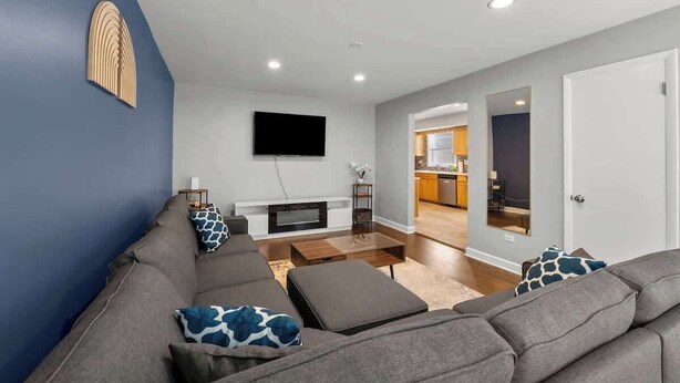 A modern living room with a grey sofa, a flat-screen TV mounted on the wall, and a wooden coffee table. - Cozy, Sunny, and Clean Apt 3 bd Free Parking (Berwyn)