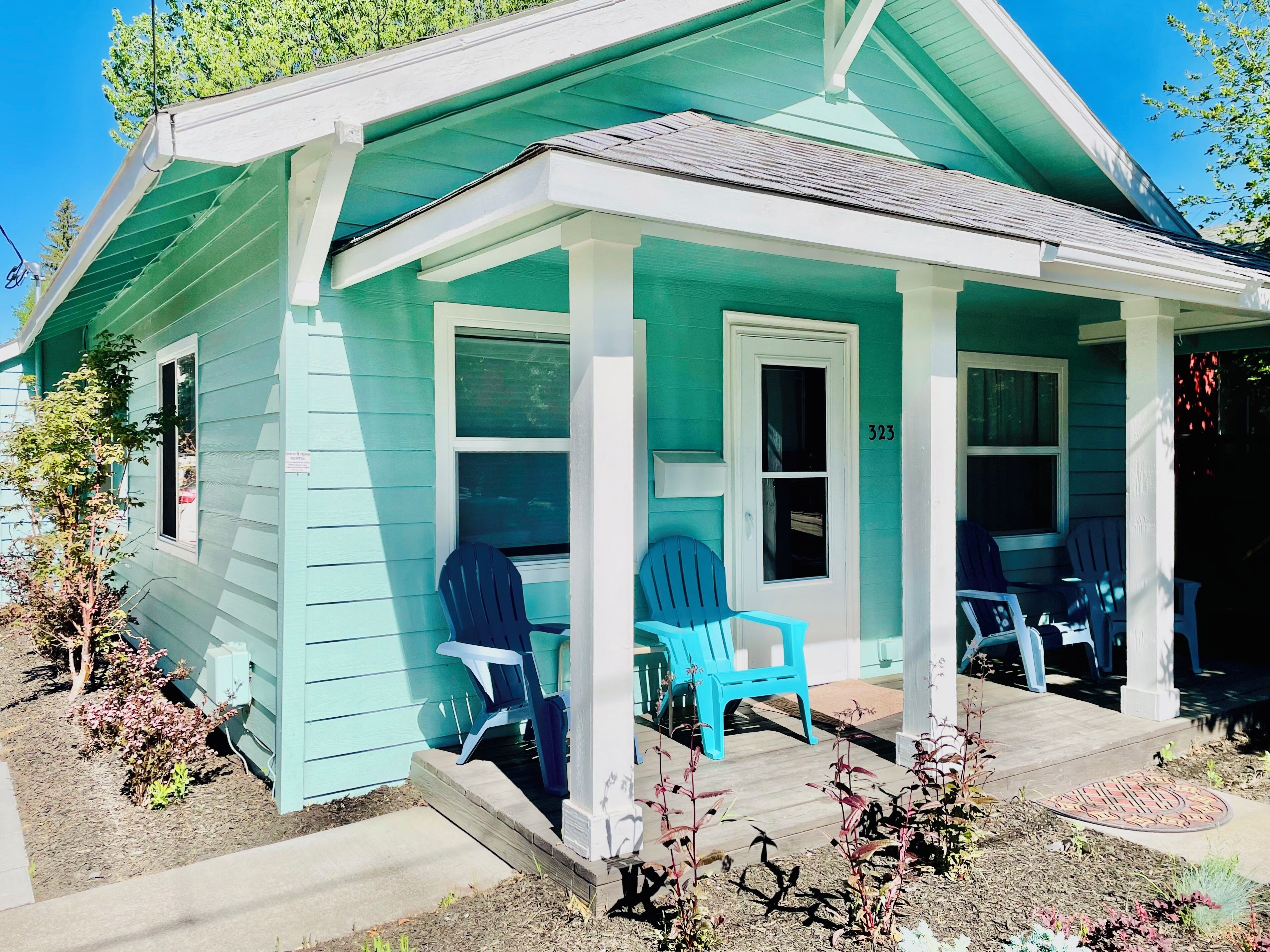 Un piccolo edificio a un piano con un portico, numero 323, con due poltrone Adirondack blu e un piccolo giardino.