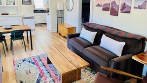 Uma sala de estar moderna com uma mesa de centro de madeira, um sofá e um tapete colorido. Há uma área de cozinha com armários e uma geladeira ao fundo.