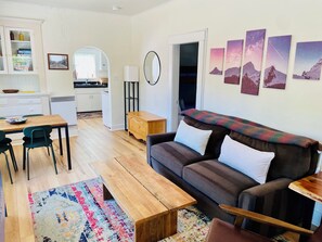 A modern living room with a wooden coffee table, a sofa, and a colorful rug. There is a kitchen area with cabinets and a refrigerator in the background.