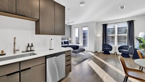 A modern kitchen with a dishwasher, a sink, and a countertop. There is a dining area with a table and chairs, and a window with curtains. - Cozy Flat w/ Pool, Gym, & Stunning City Views (Birmingham)