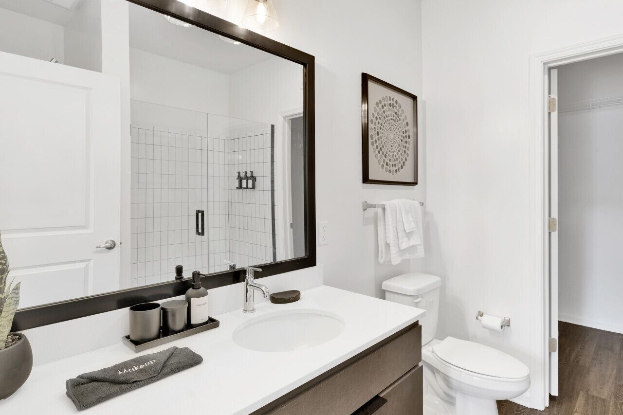 Un bany modern amb un mirall gran, un lavabo blanc i un vàter. Hi ha una obra d'art amb marc a la paret i un penjallatot.