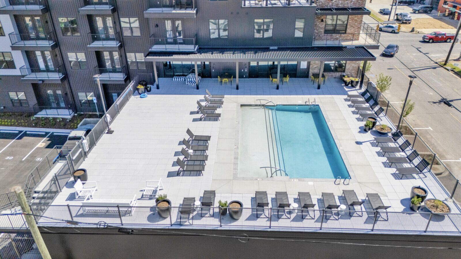 A rooftop pool area with sun loungers and a swimming pool, surrounded by a fence and a building with balconies.