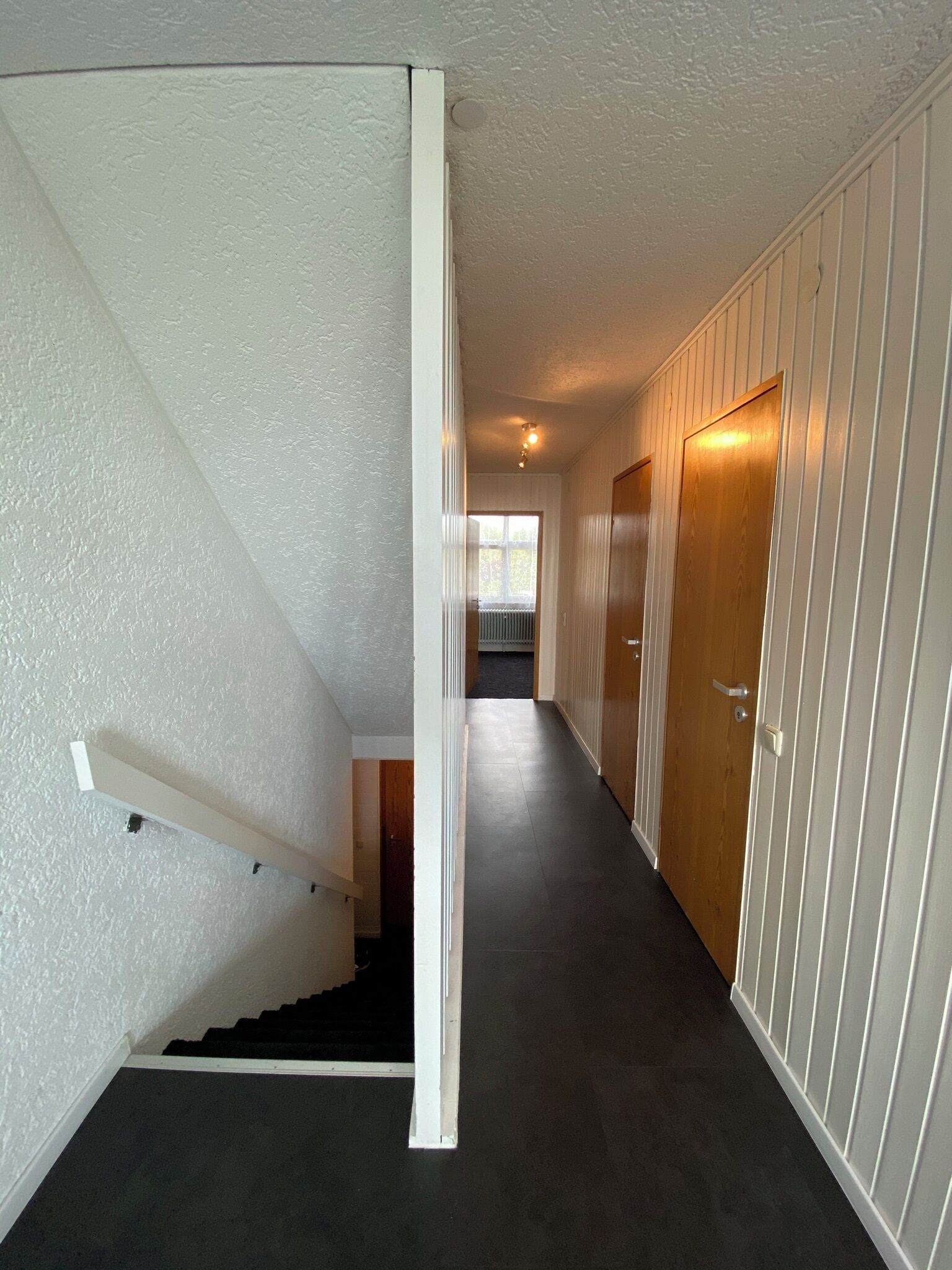 Ein schmaler Flur mit dunklem Boden, weißen Wänden und einer Holztür am Ende.