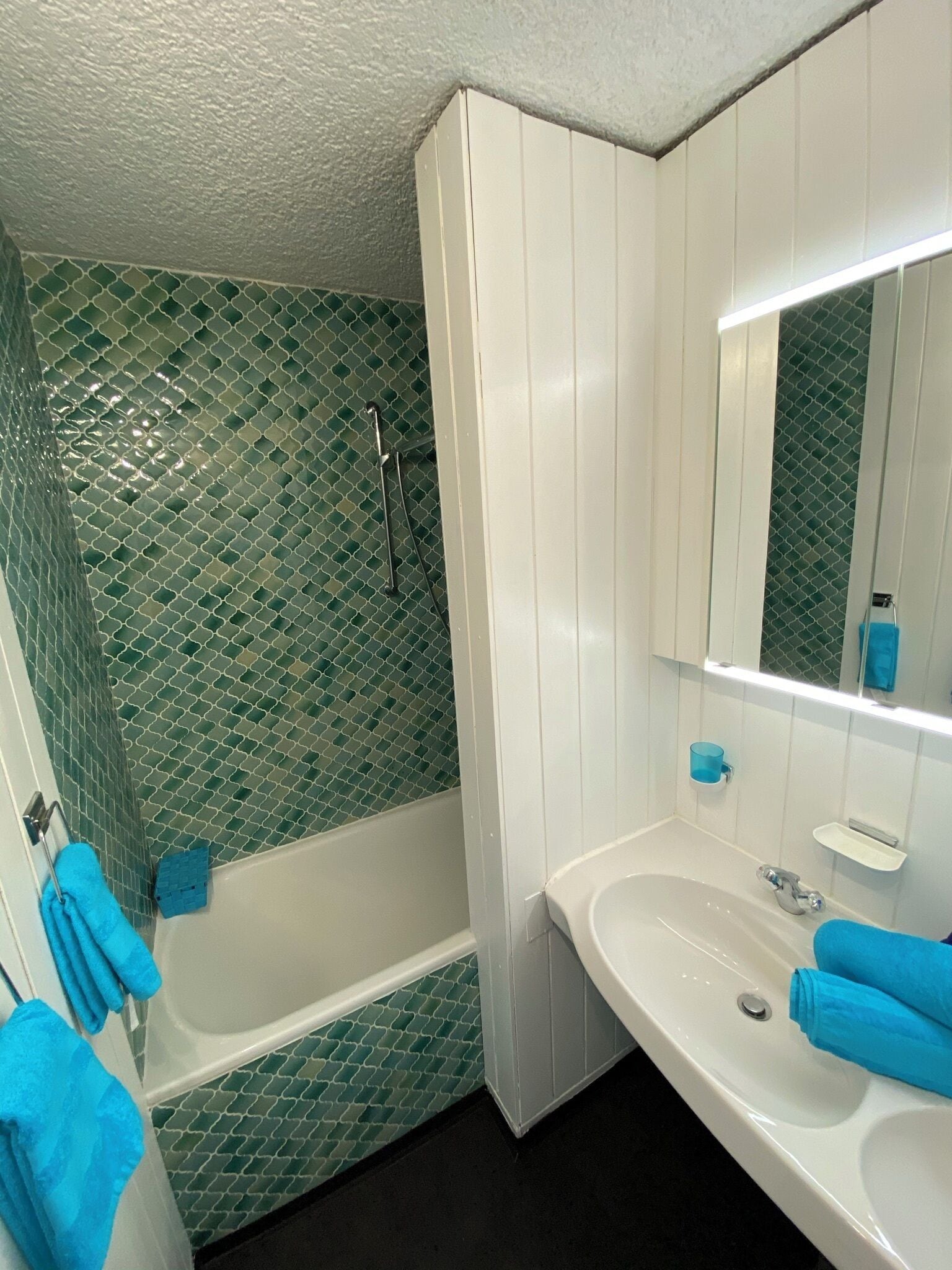 A bathroom with a green mosaic tile wall, a white bathtub, a white sink with a mirror, and blue towels.