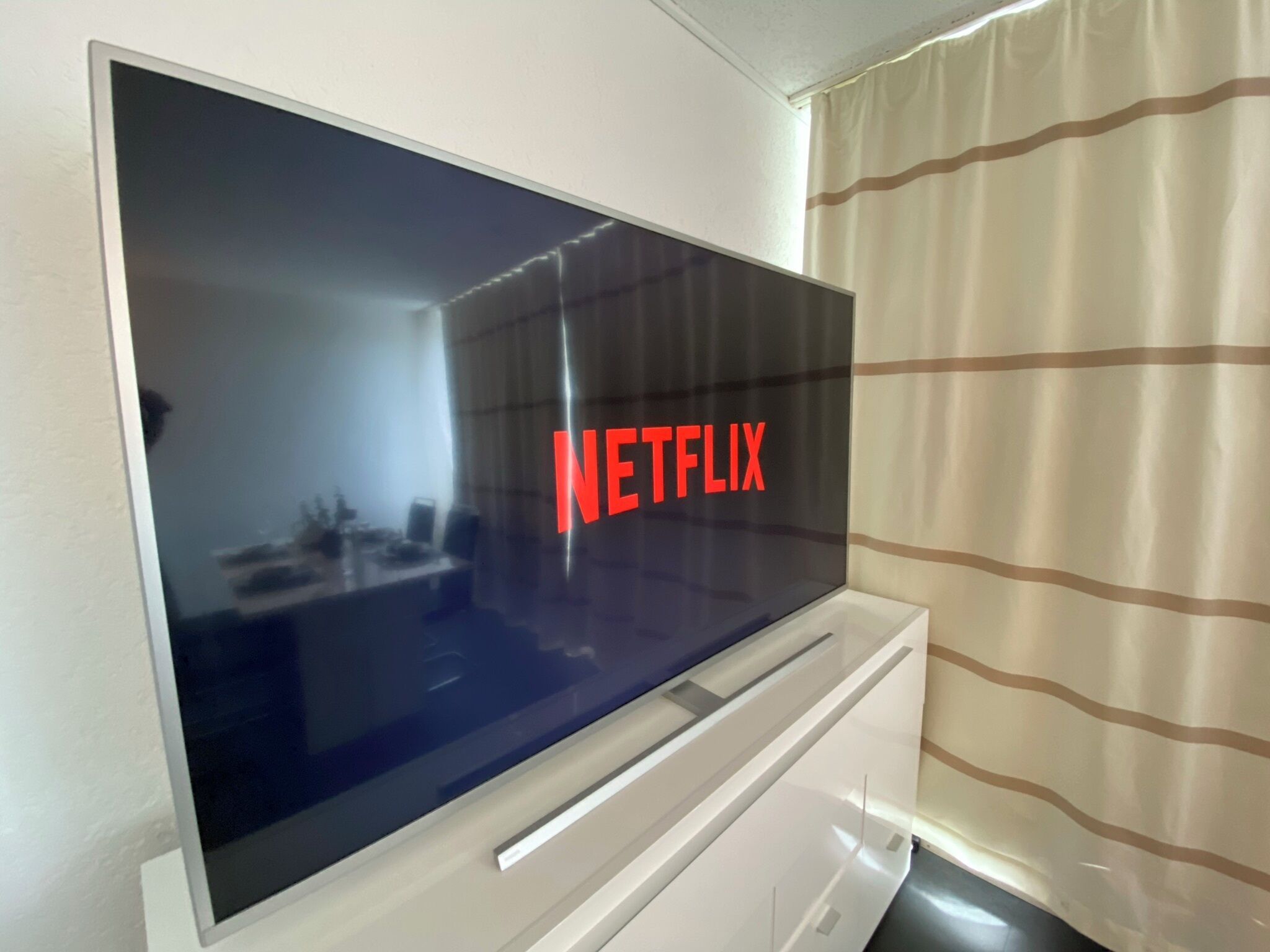A flat-screen TV mounted on a white cabinet with a reflection of a room and a window with curtains.
