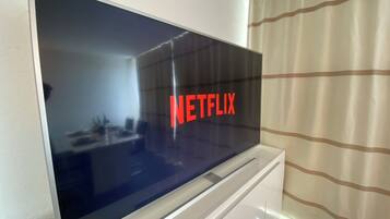 A flat-screen TV mounted on a white cabinet with a reflection of a room and a window with curtains.