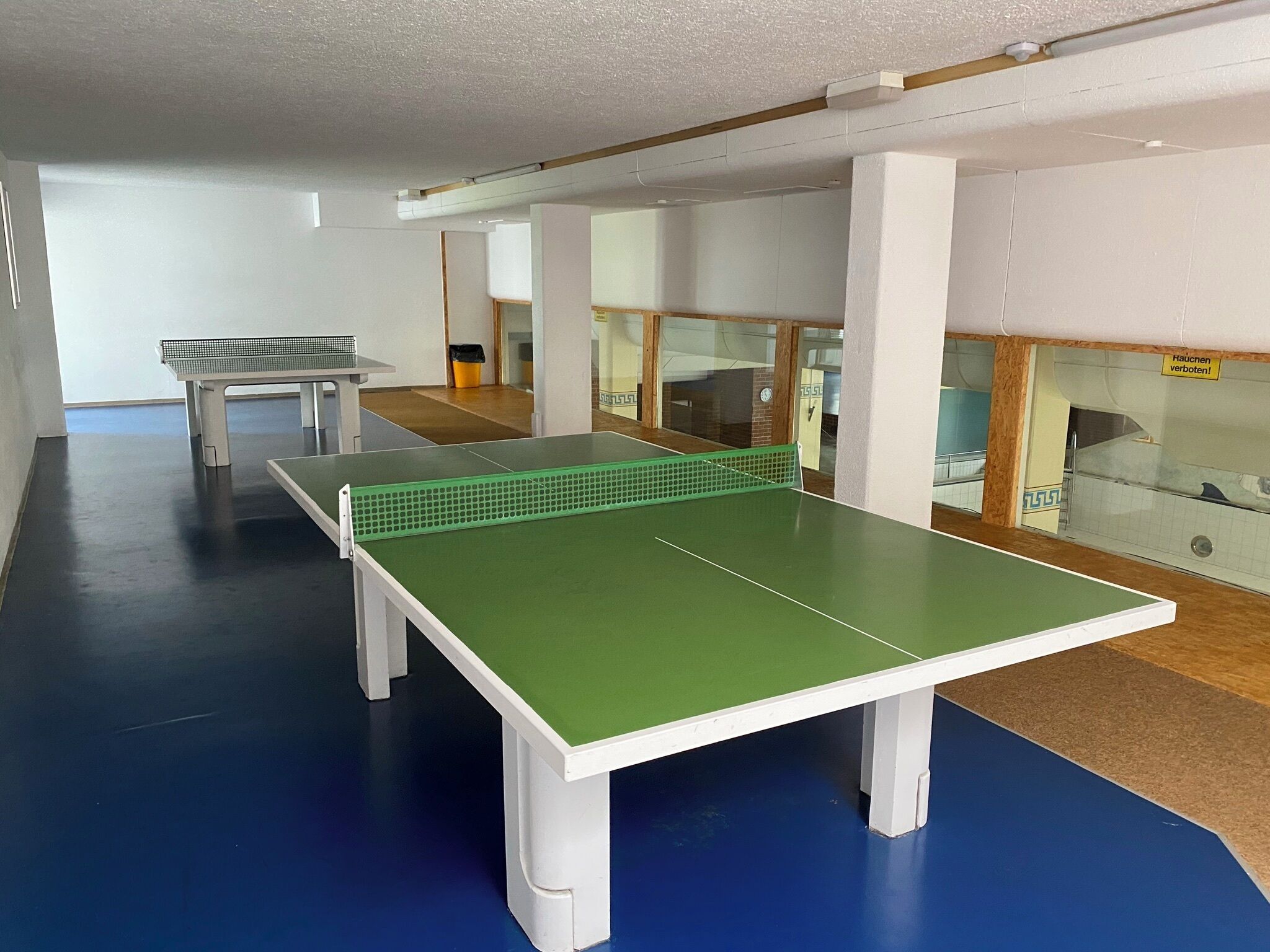 A ping pong table in a room with a blue floor, white walls, and a small table in the background.