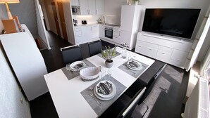 A modern kitchen with white cabinets, a dining table set for four, and a large flat-screen TV on the right. - COZY PENTHOUSE in the middle of the BAVARIAN FOREST +NETFLIX +POOL +SAUNA +GYM (Sankt Englmar)