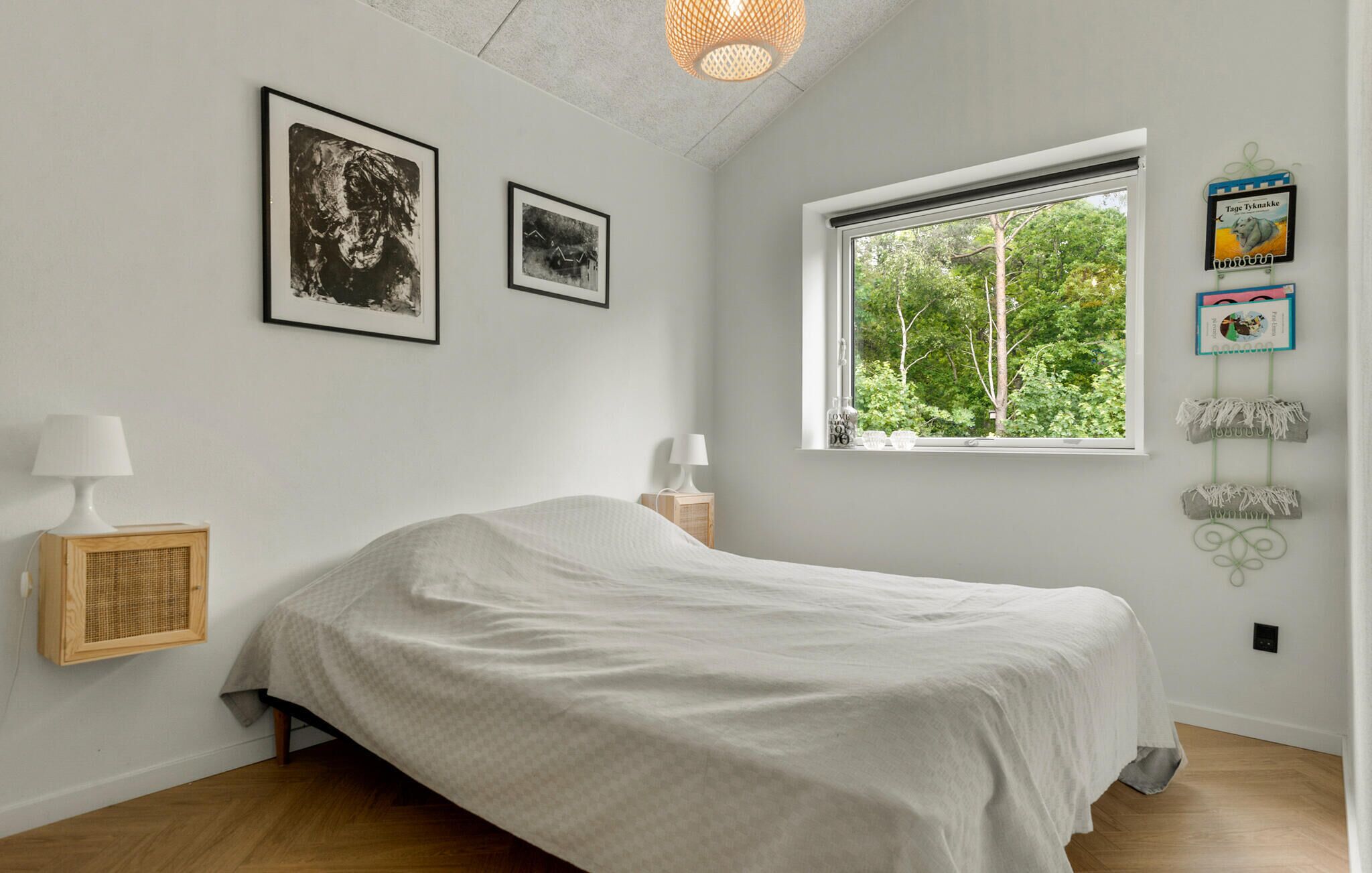 A bedroom with a bed, two framed pictures, a window with a view of trees, a lamp, and a small wooden cabinet.