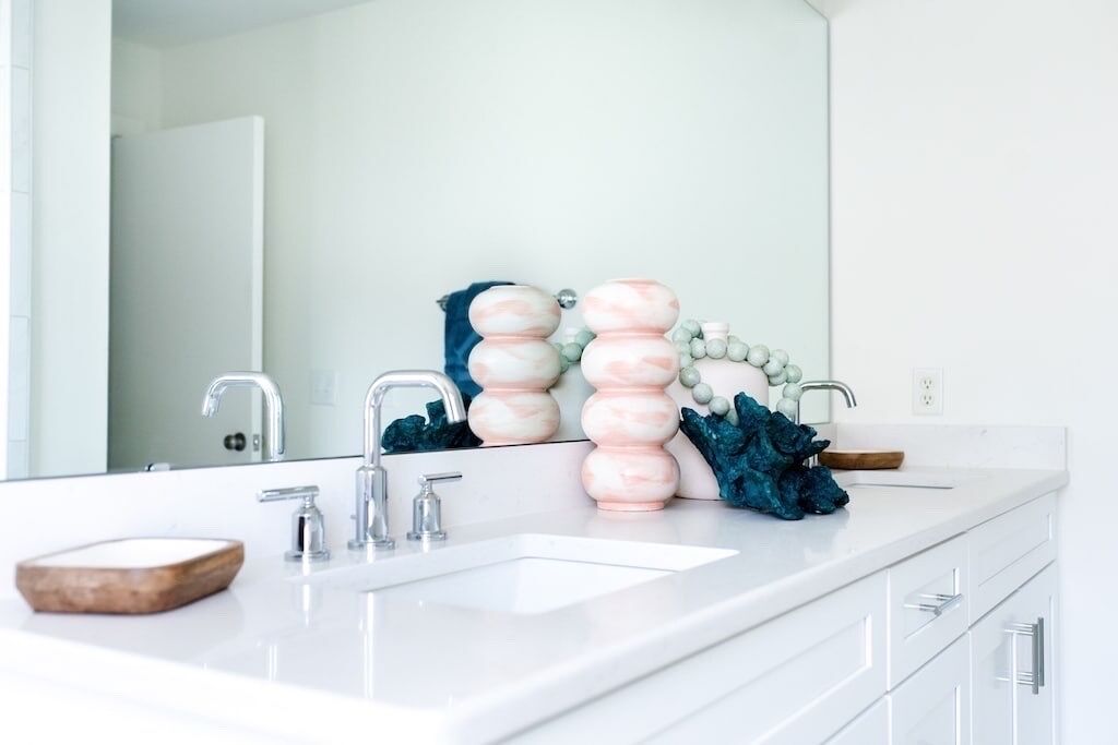 Un baño moderno con un lavamanos blanco, dos grifos, un espejo y objetos decorativos, incluyendo un cuenco de madera y una escultura.