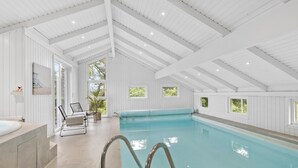 Una piscina interna con soffitto alto, ampie finestre e un'area relax con sedie e un tavolo.