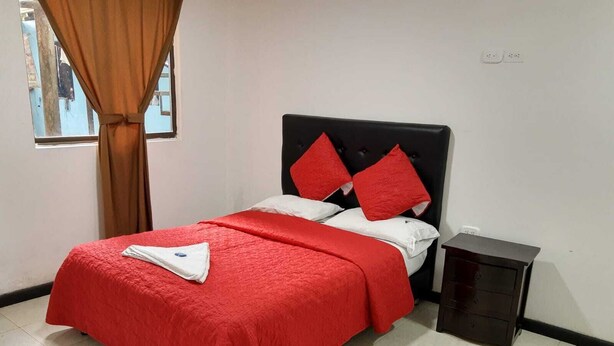 Ein Bett mit roter Bettdecke und Kissen, ein kleiner Nachttisch und ein Fenster mit Vorhängen.