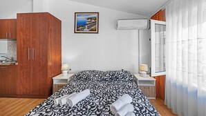 A bedroom with a bed, wooden wardrobe, a painting, an air conditioning unit, and a window with curtains.