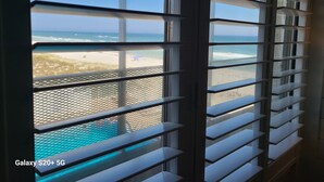 Ein Fenster mit horizontalen Jalousien, Blick auf den Strand und einen Pool.