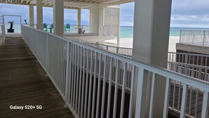 Eine überdachte Holzterrasse mit weißen Geländern führt zu einem Strand mit türkisblauem Wasser und weißem Sand.