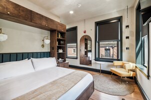 A modern bedroom with a large bed, a wooden headboard, a small round table, a chair, and a view of the outside.
