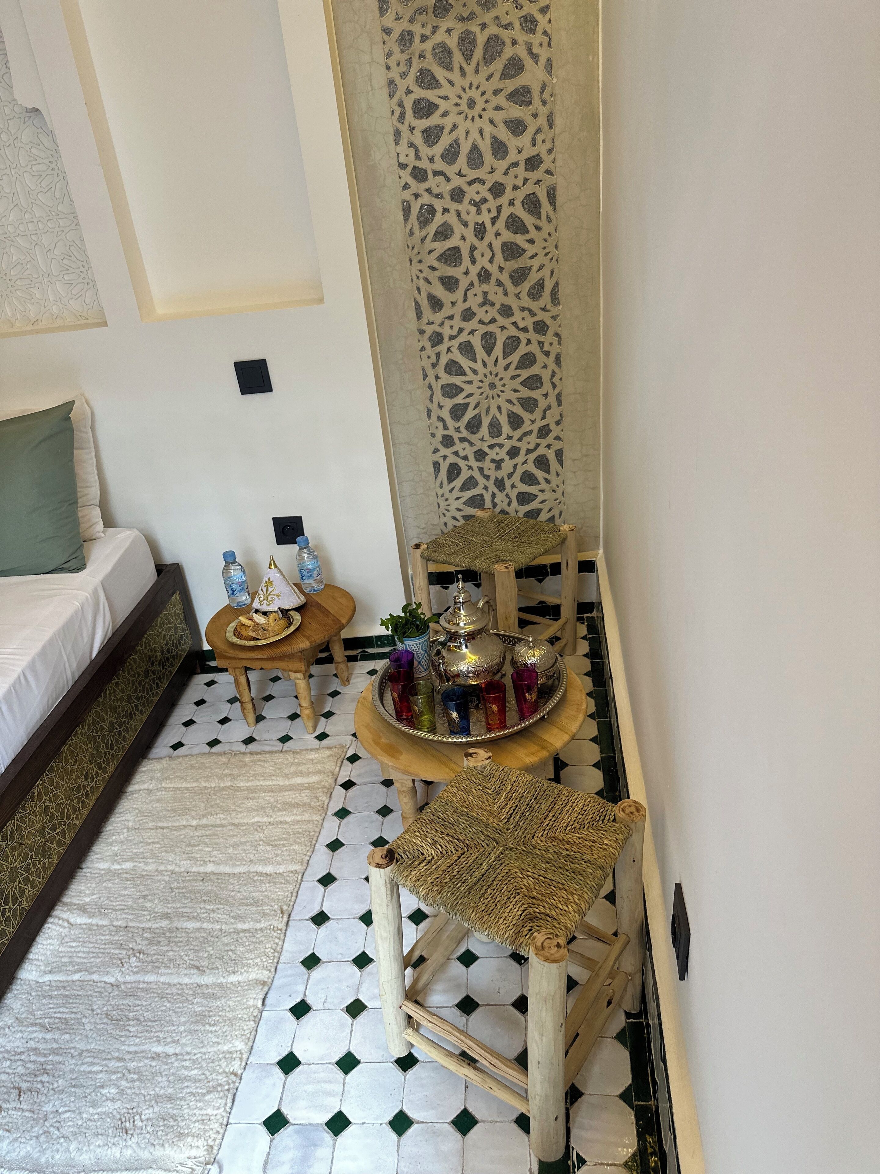 A cozy room with a bed, a small table with a tray of drinks and a teapot, a decorative wall panel, and a woven chair.