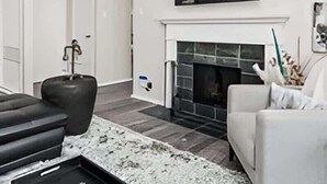 Un salon moderne avec une cheminée, un téléviseur à écran plat fixé au-dessus, un grand pouf noir et un canapé blanc.
