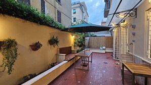 A narrow courtyard with a wooden bench, a table, and an umbrella. There are potted plants on the walls and a wooden fence in the background. - 2 bedroom apartment in rome (Roma)