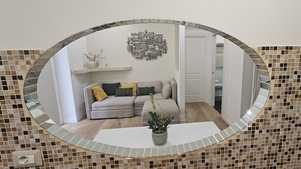A room with a tiled floor, a white sofa with cushions, a small table with a potted plant, and a wall-mounted mirror. - 2 bedroom apartment in rome (Roma)