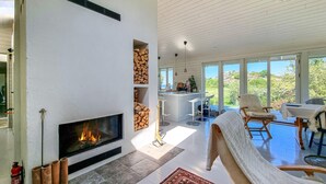 A modern living room with a fireplace, a kitchen area, and a view of the outdoors.