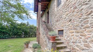 Un edificio de piedra con una escalera de madera que lleva a una ventana. Hay plantas en macetas en los escalones y un área con césped y flores amarillas.
