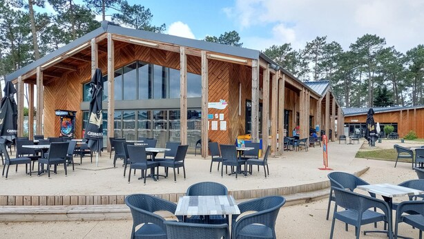 Un espace extérieur aménagé avec des tables et des chaises, une structure en bois dotée de grandes fenêtres et une enseigne sur laquelle on peut lire « Grimmer ».