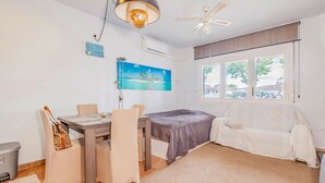 Une pièce comprenant une table à manger, des chaises, un lit recouvert d’une couverture blanche, un ventilateur de plafond, un appareil de climatisation, un tableau représentant une plage et une fenêtre donnant sur des maisons et des arbres.