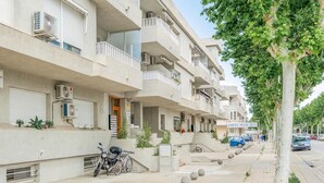 Un immeuble résidentiel à plusieurs étages, doté de balcons, de climatiseurs et offrant une vue sur la rue, où l’on peut voir des voitures garées et un arbre.