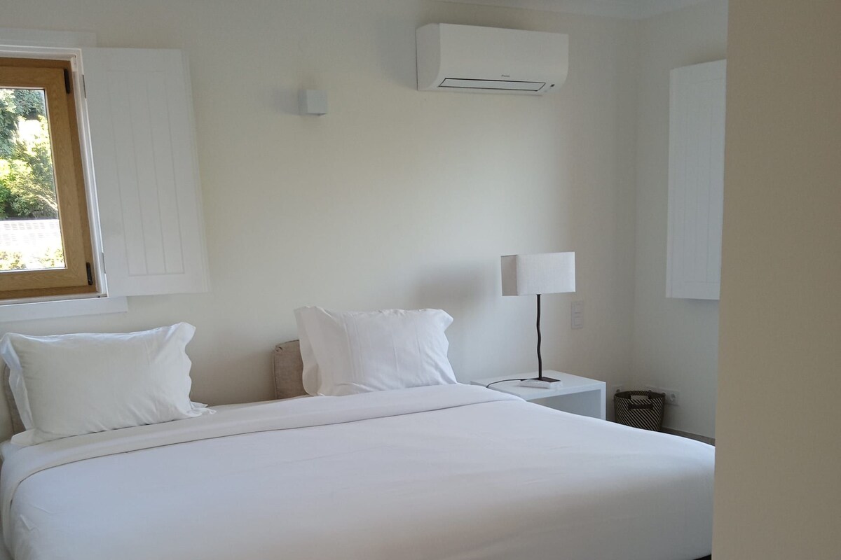 A bedroom with a large bed, a bedside table with a lamp, an air conditioning unit on the wall, and a window.