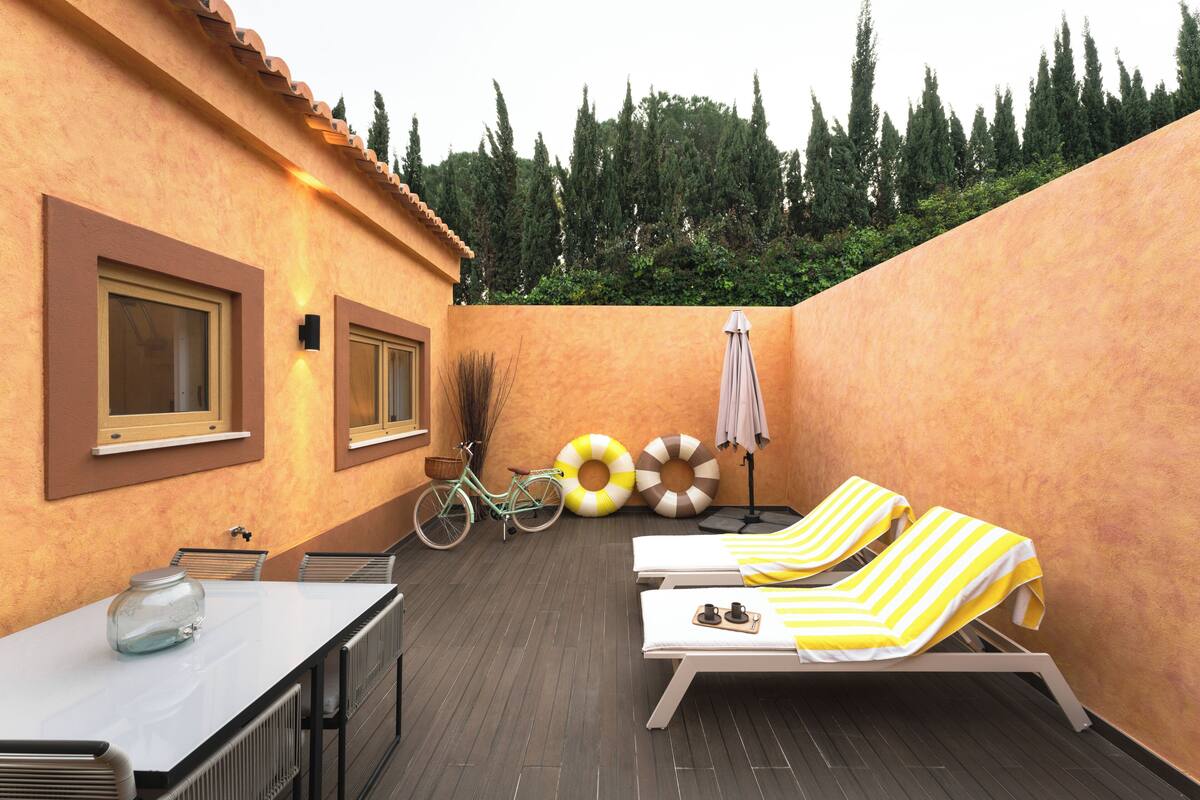 A sunlit patio with a table, two lounge chairs, a bicycle, and a wall with windows.
