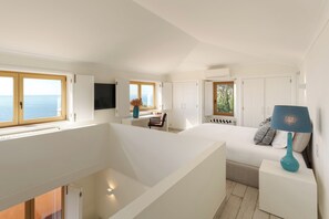 A modern interior with a large white bed, a blue lamp, and a view of the sea through large windows.