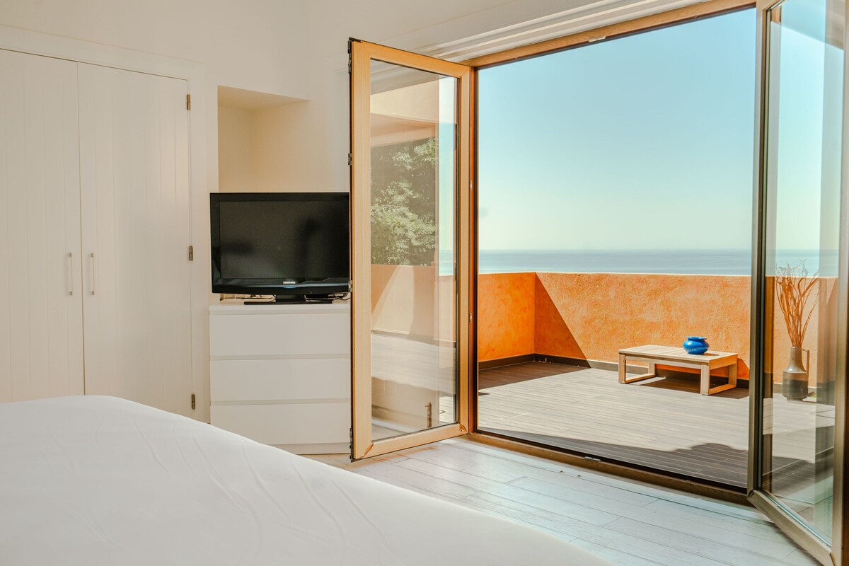 A modern bedroom with a large bed, a television, and a sliding glass door leading to a balcony with a view of the sea.