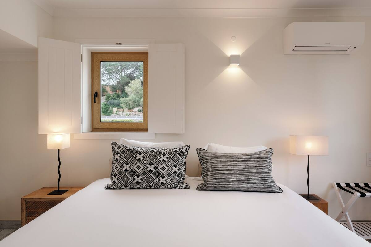 A bedroom with a bed, two bedside lamps, a window with a view, and an air conditioning unit.