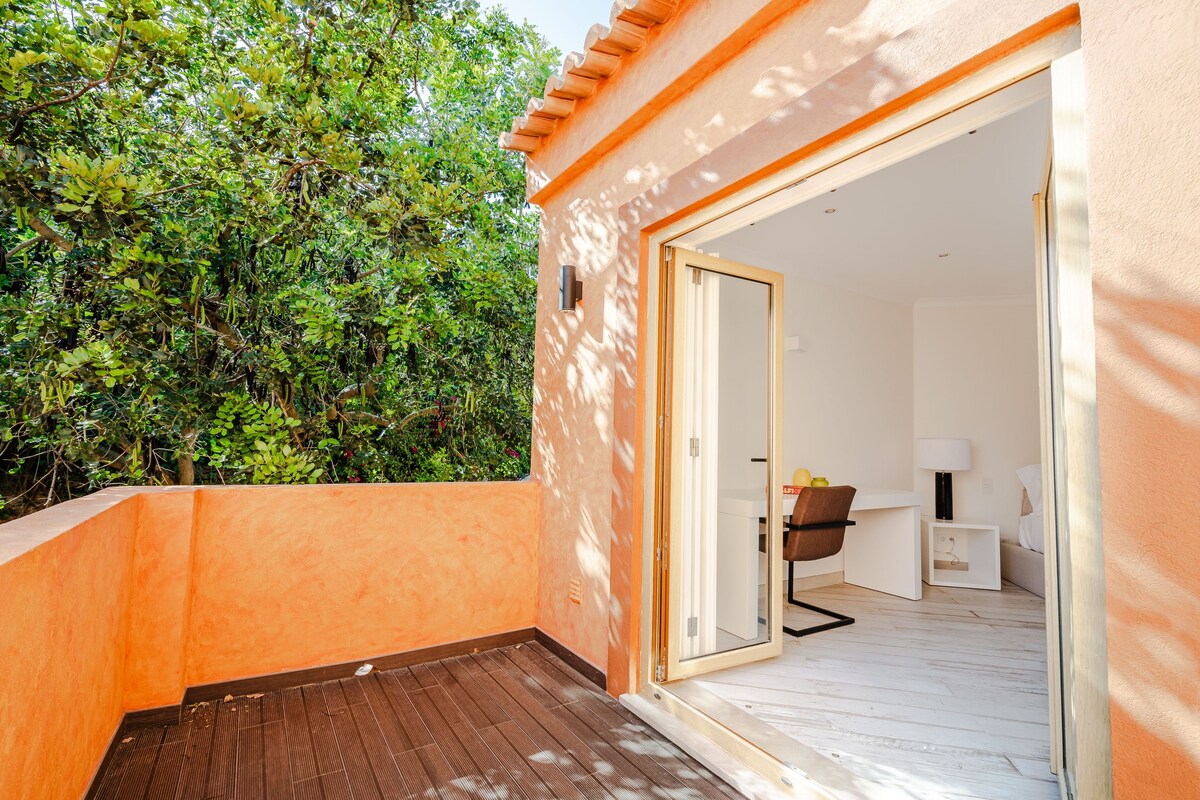 A balcony with a wooden floor, a glass door leading to a room with a desk, chair, and a small table with a lamp.