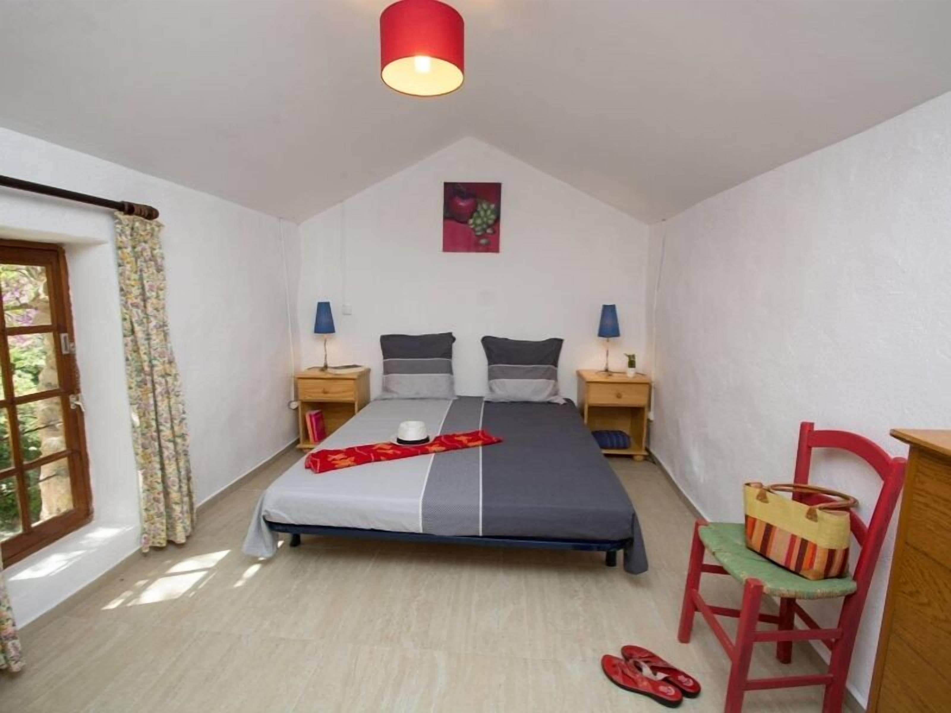 Una camera da letto con un letto, due comodini, una sedia, una lampada rossa e una finestra con tende.