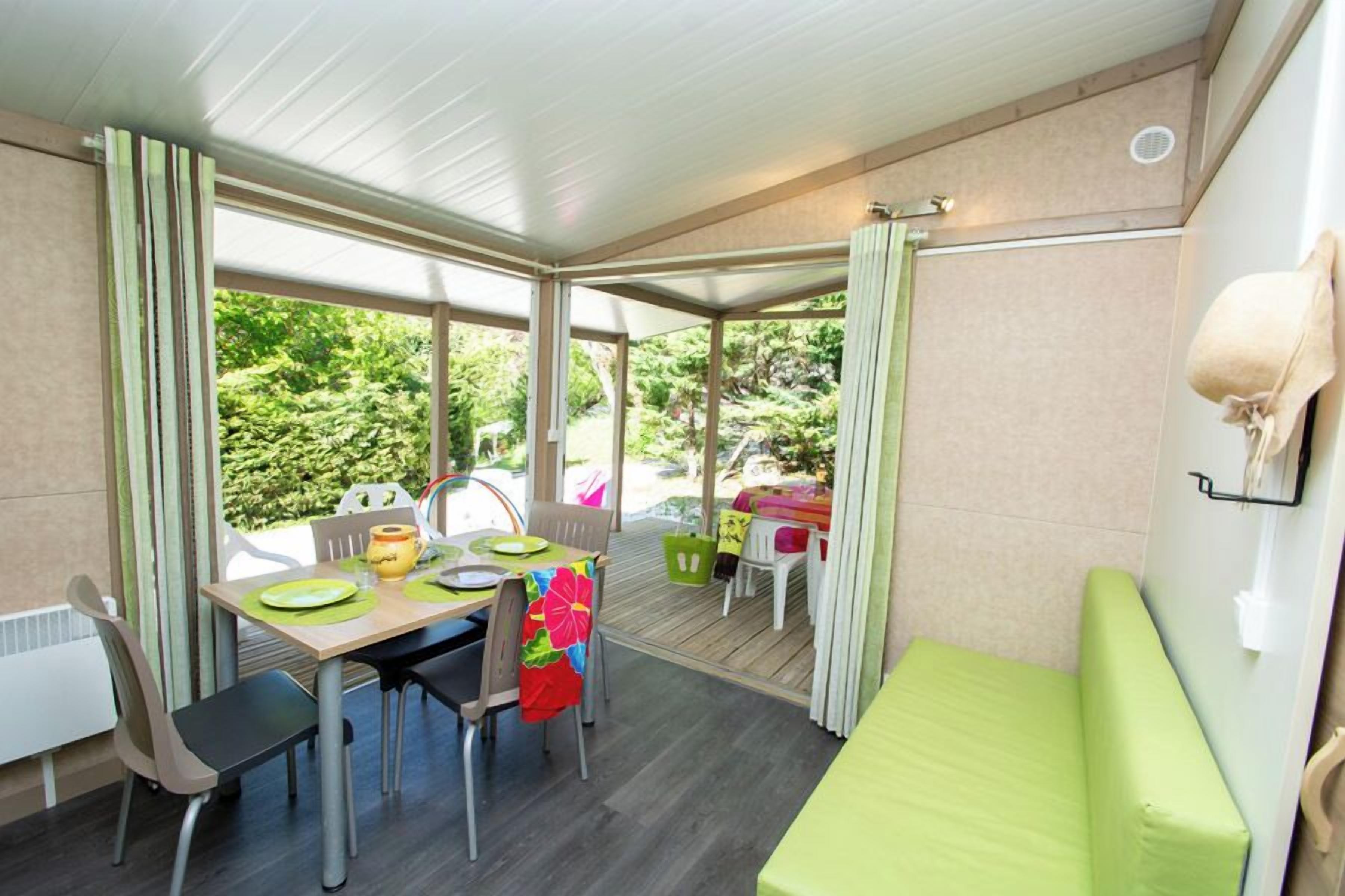 Comedor con una mesa preparada para dos personas, un banco acolchado verde y vistas al exterior con vegetación.
