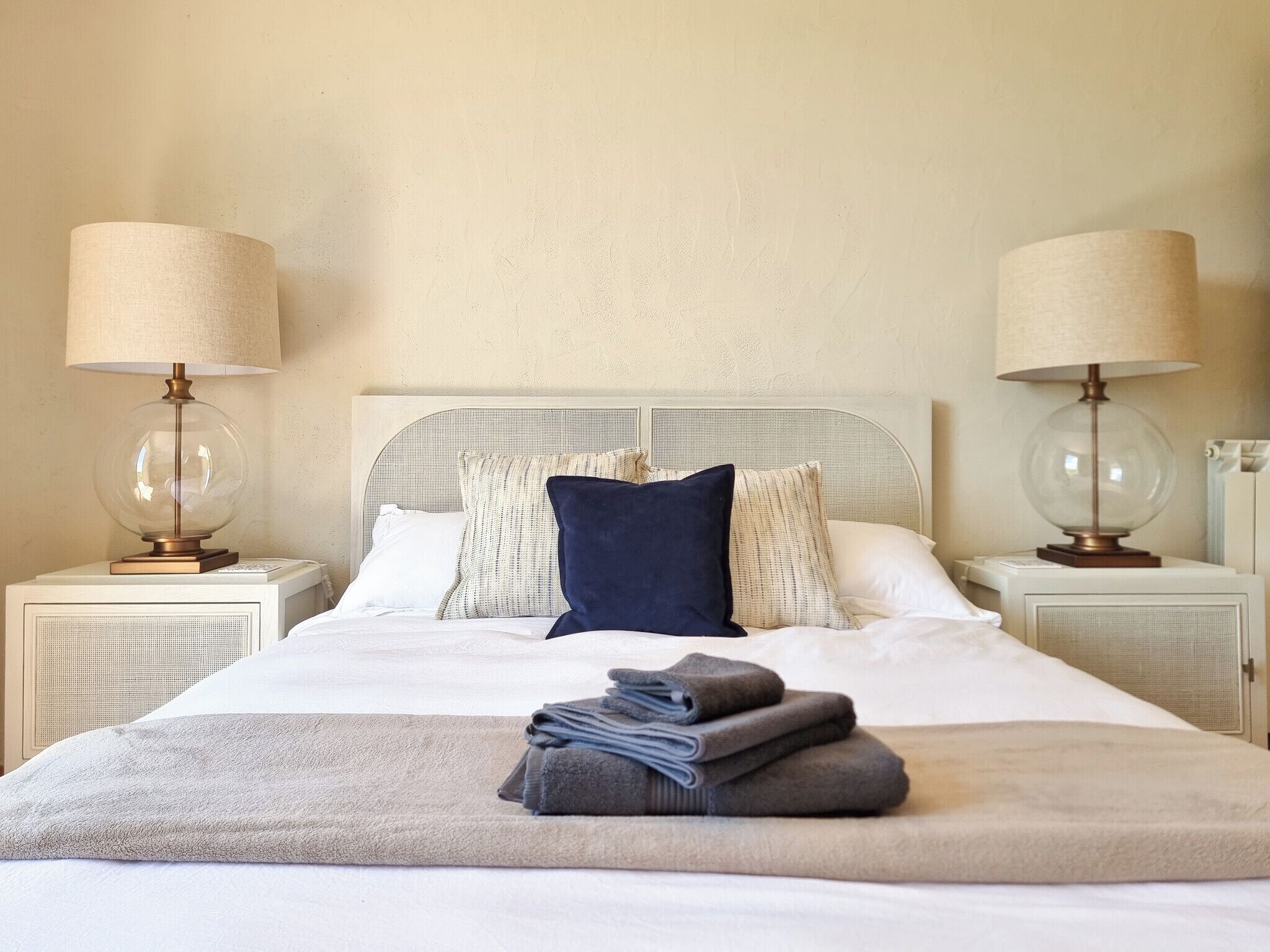 A neatly made bed with a white and gray duvet, two lamps on either side, and a stack of folded towels on the bed.