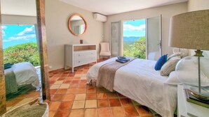 Une chambre à coucher comprenant un lit, une commode, un miroir, une chaise et donnant sur l’extérieur.
