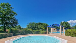 Ein rechteckiger Swimmingpool mit einer gefliesten Poolterrasse, einer Pergola mit blauen Holzbalken und einer Steinmauer im Hintergrund.