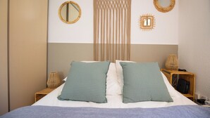 Ein Bett mit zwei Kissen, ein Nachttisch mit Lampe sowie Wanddekorationen, darunter ein Wandbehang aus Makramee und zwei Spiegel.