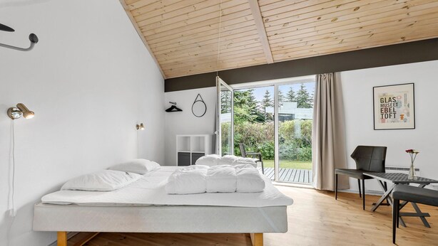 Ein modernes Schlafzimmer mit einem großen Bett, einem Schreibtisch, einem Stuhl, einer Holzbalkendecke und Blick ins Freie.