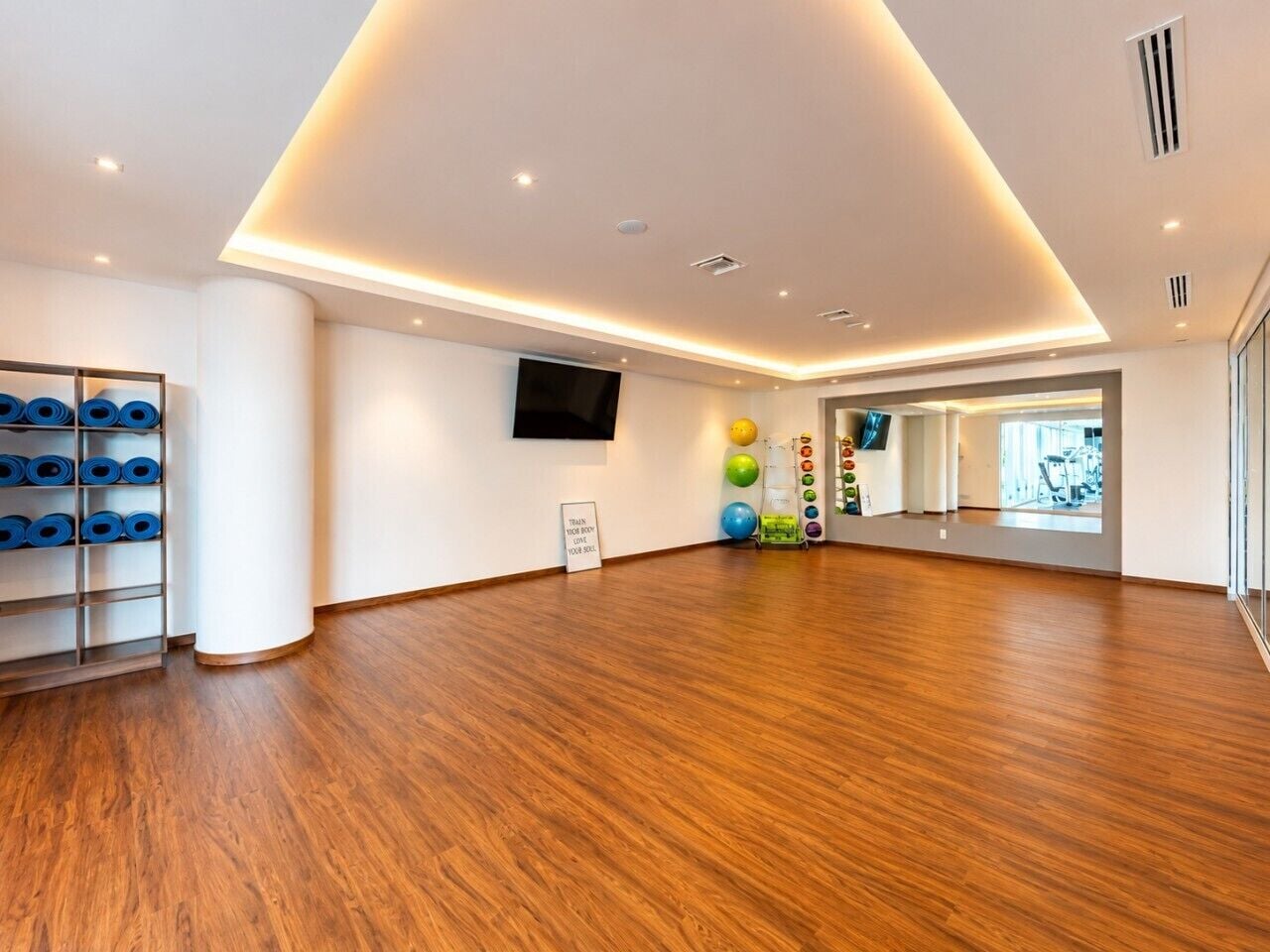 Quarto espaçoso com piso de madeira, televisão fixada na parede e um suporte com tapetes de exercício azuis.