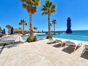 Ein Poolbereich mit Liegestühlen und Sonnenschirmen, Palmen und freiem Blick auf den Ozean.
