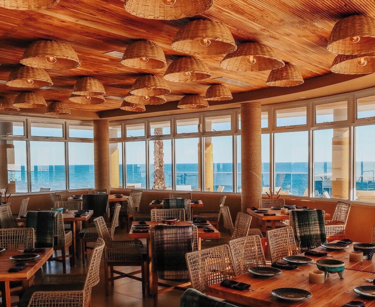 Een restaurant met rieten meubels, grote ramen en uitzicht op zee.