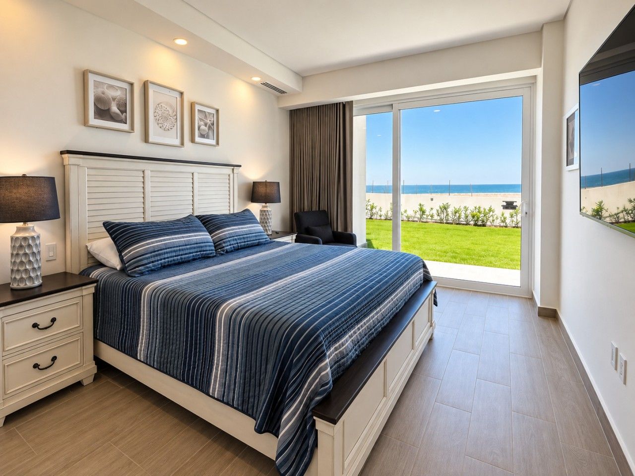 Een slaapkamer met een groot bed, een nachtkastje, een lamp en uitzicht op het strand door een schuifdeur.