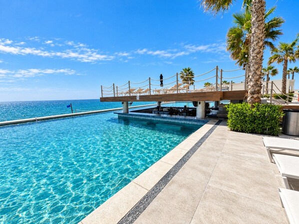 A clear blue pool with a wooden deck and lounge chairs, palm trees, and a view of the ocean.